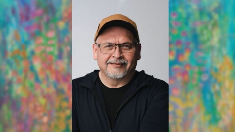 A picture of a man wearing an orange hat with gray facial hair with a painting with blue yellow and pink colors behind him.