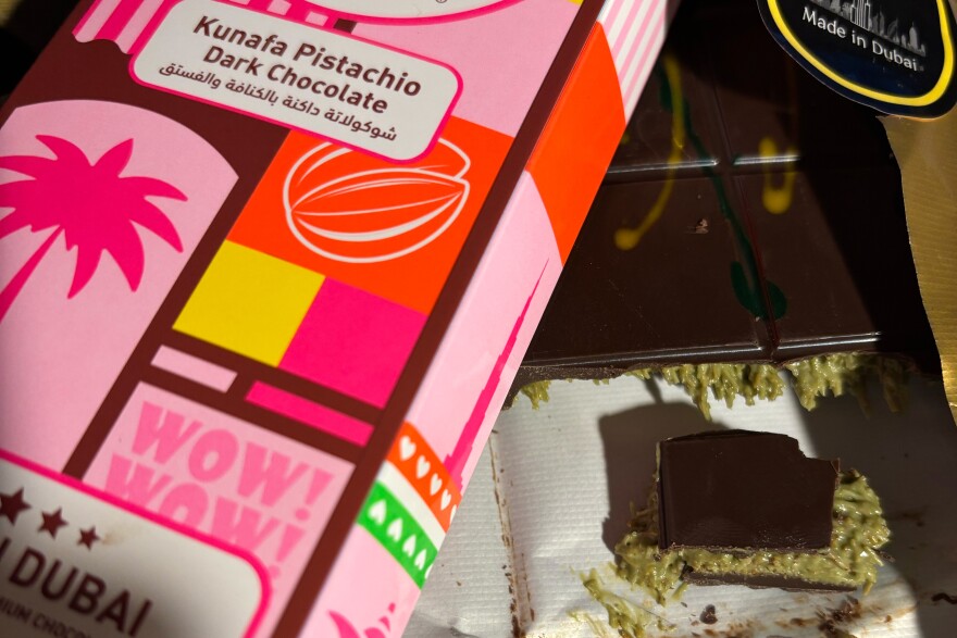 Pieces of a chocolate bar with green paste inside it, and part of the bright, pastel-colored packaging crossing over it on top. The word Dubai in the bottom left corner and "made in Dubai" in the top right.