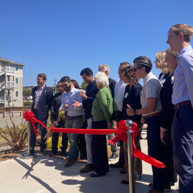 A person wearing a button-down shirt and sunglasses cuts a red ribbon alongside several other people.