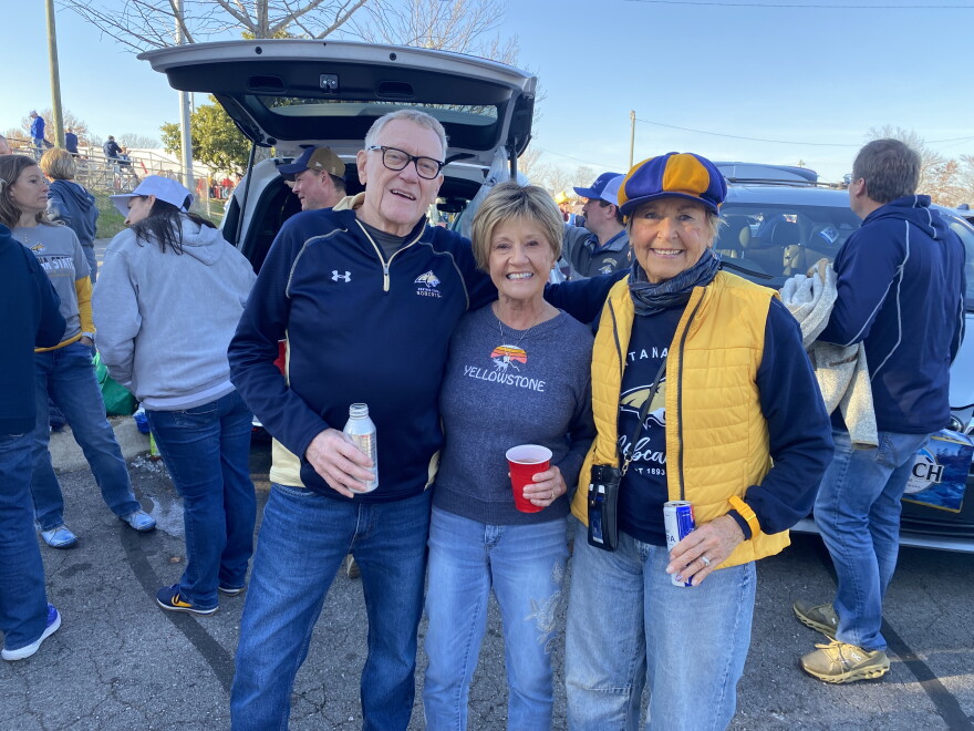 Three smiling adults pose together at a tailgate party. The man and two women wear casual clothes with sports logos. They stand in front of an open SUV, holding drinks, with other fans in the background.