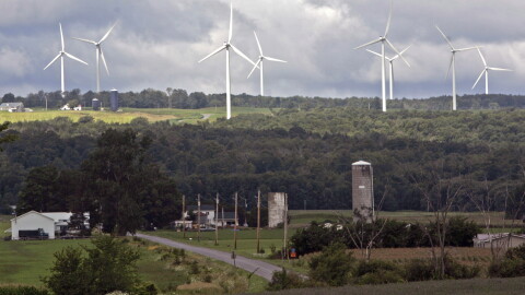 A rural landscape. Farms and trees are visible in the foreground. In the background are 8 large white windmills.
