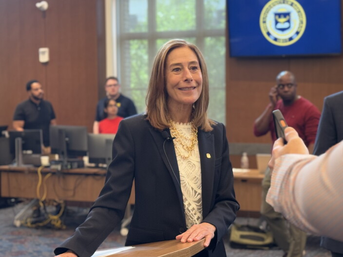 C. Cybele Raver, wearing a navy blazer and white lace top with a gold chain necklace, stands smiling at a wooden lectern during a media interview. In the background, a monitor displays the seal of the University of Michigan.