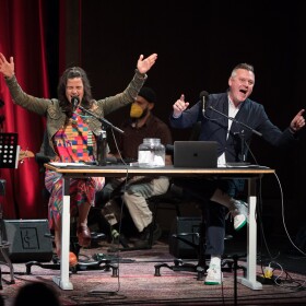 Two podcast hosts on the stage in front of their microphones. 