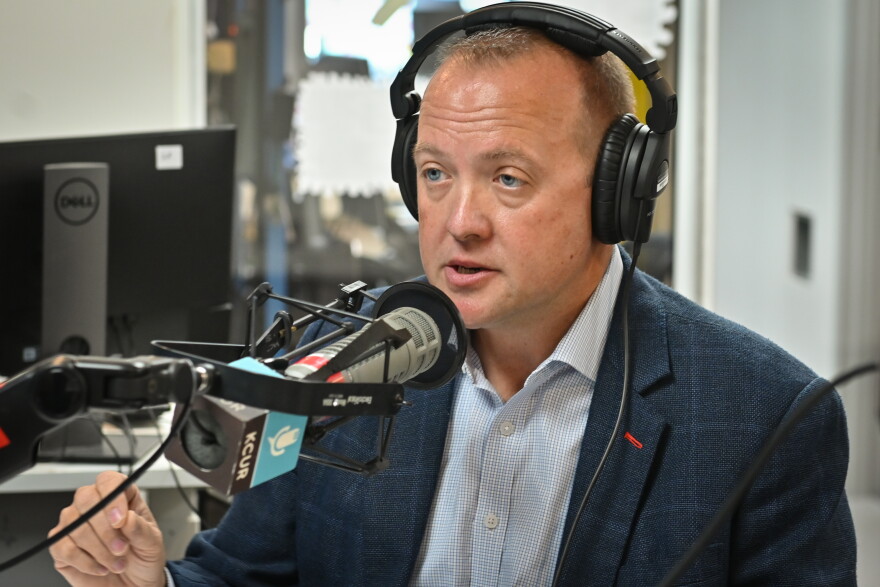 A man wearing a blue sports coat  is wearing a set of headphones, sitting inside a radio studio. He's talking and gesturing with his right hand.