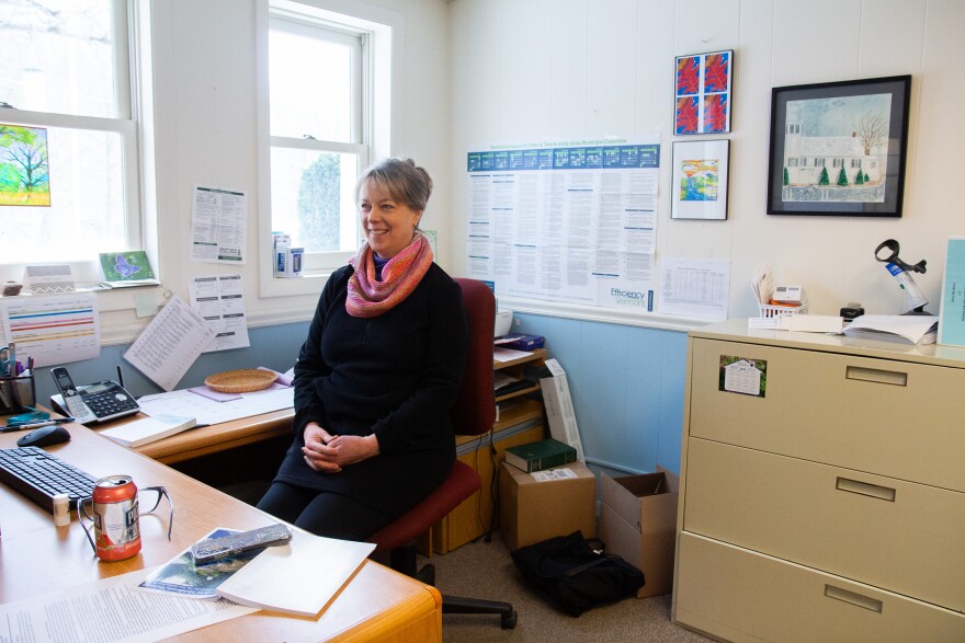 A photo of a person wearing a black dress and tights and a pink and purple scarf. Her hair is greyish blonde and is up in a bun. Around her is a desk with a keyboard, glasses and drink, and a painting of a white town hall. More papers and boxes and cabinets are around the room, which has natural light coming in through the windows.