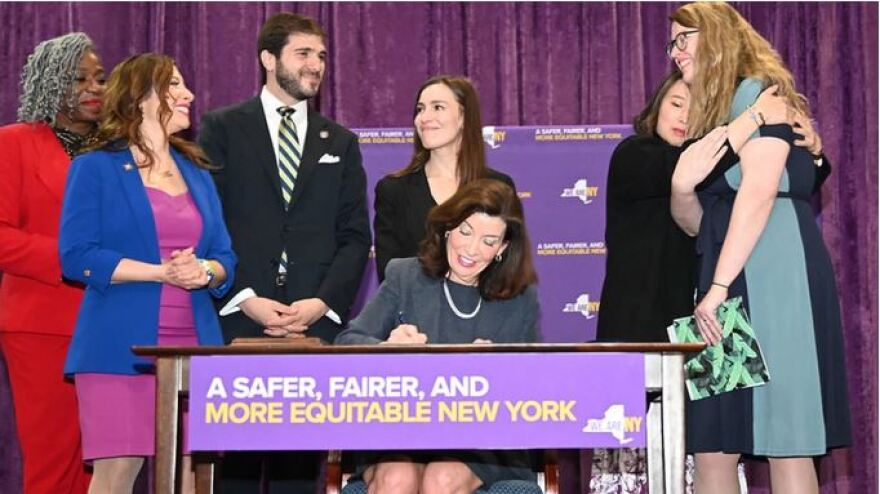 Gov. Kathy Hochul, seated, signs the legislation as a group stands around her.