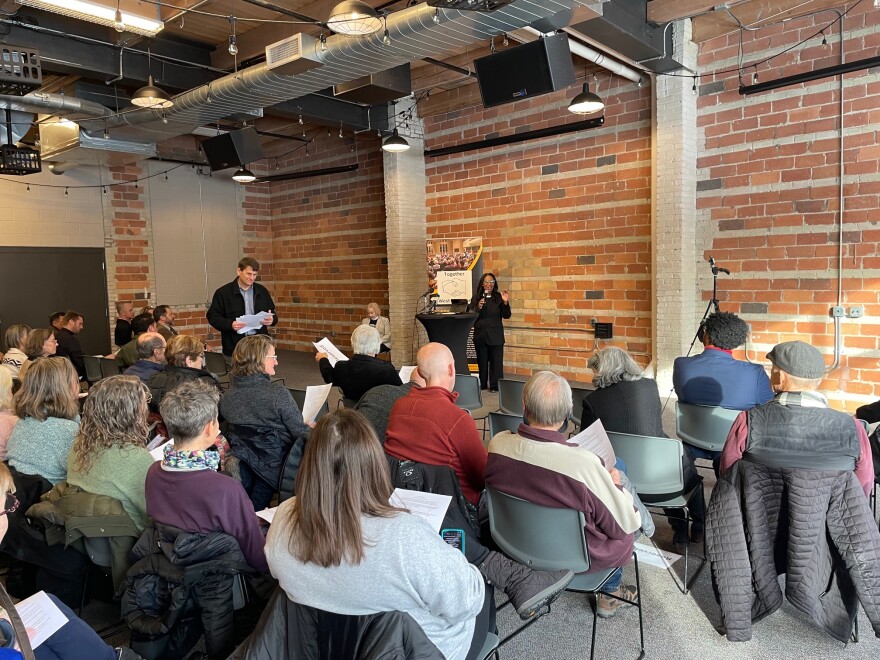People sit in chairs facing a speaker at a podium. A brick wall is behind.