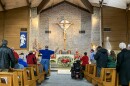 Congregants stand and sit during worship as part of a inclusive Christmas Mass at St. Bernadette Catholic Church. There is a wheelchair user in the isle near the front, facing an altar decorated with Christmas wreaths and lights.