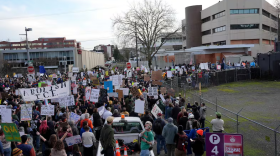 A crowd of people with signs stand along a fenced area.