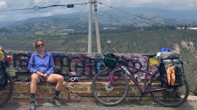 Smiling woman in shorts sitting on bench with graffiti. Two biked leaning against the bench filled with rucksacks. Mountains in background