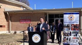 A man in a navy suit jacket speaks at a podium with the North East ISD emblem. Four women and a man in business attire stand behind him. Behind them is a school building with a banner that reads "Wilshire Safety Training Center."