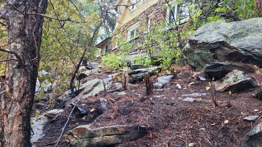 Burned ground on a steep mountainside is seen ten feet from a house. Char marks are on the rocks, and the forest floor, and burned pine needles are also visible. A small amount of green, unburned flora sits between the charred area and the house.