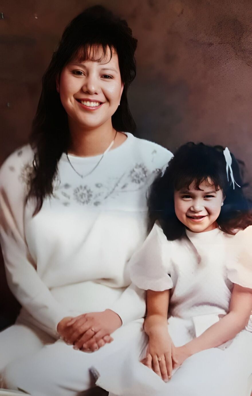 A mother and daughter are sitting side by side with big smiles on their faces. They are wearing all white/cream clothing. They have dark eyes and hair and are pretty.