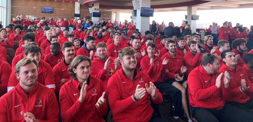 Large group of young men all wearing red jackets and applauding as they look up to a television monitor