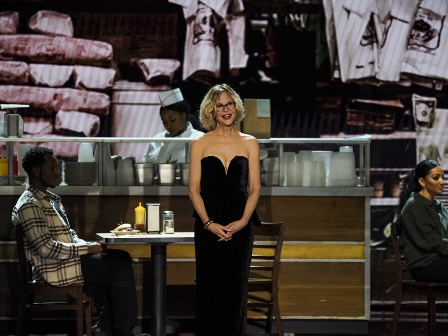 A woman wearing a black formal gown speaks on stage at a theater with a diner scene in the background.