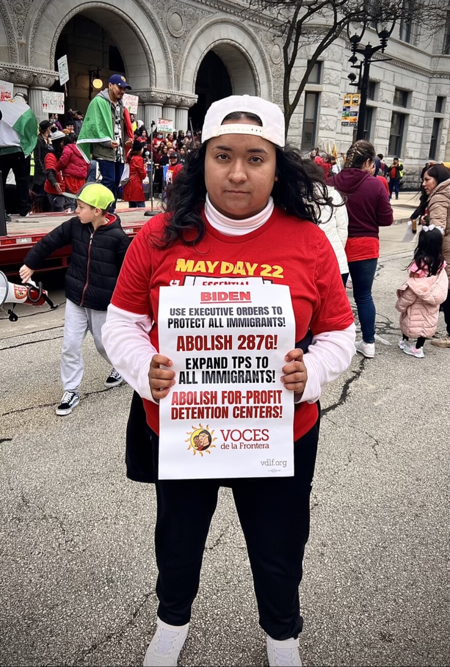 A woman with dark curly hair looks seriously at the camera. She holds an immigration protest sign that calls on Biden to protect all immigrants.