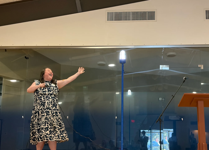 Woman in black and beige patterned dress lifts hands up to a light bulb on the stage 
