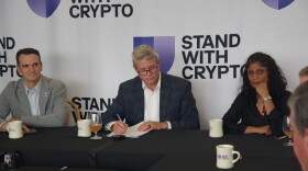 Several people sit around a black table and appear listening to a speaker who is off camera. Behind them, large white banners read "Stand with Crypto"