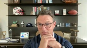 A man in glasses and navy sweater clasps hands, sitting in front of a bookcase with sports memorabilia and a dozen or so books