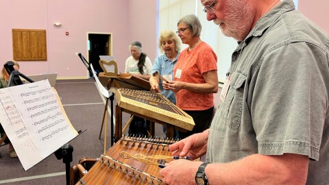 A circle of students stand in front of their hammered dulcimers, a trapezoidal string instrument which they strike with wooden mallets.