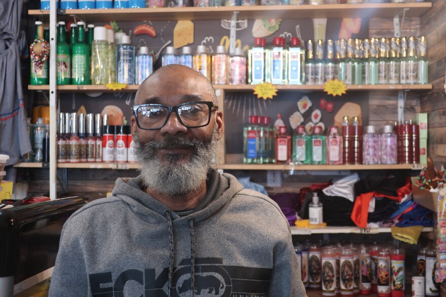 A Black man with glasses and a long grey beard looks at the camera. Behind him are bottles and candles.