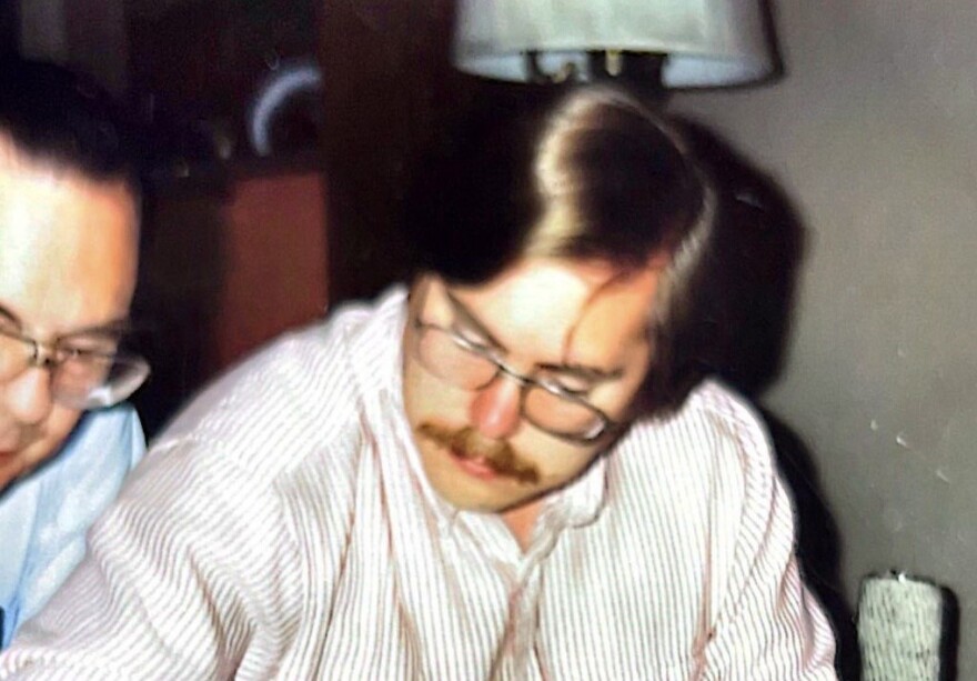 A man with light brown hair and a thick brown mustache looks down wearing 80s style glasses and a striped, collared shirt. 