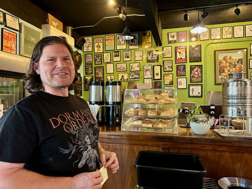 A man stands in front of a counter displaying pastries and coffee. 