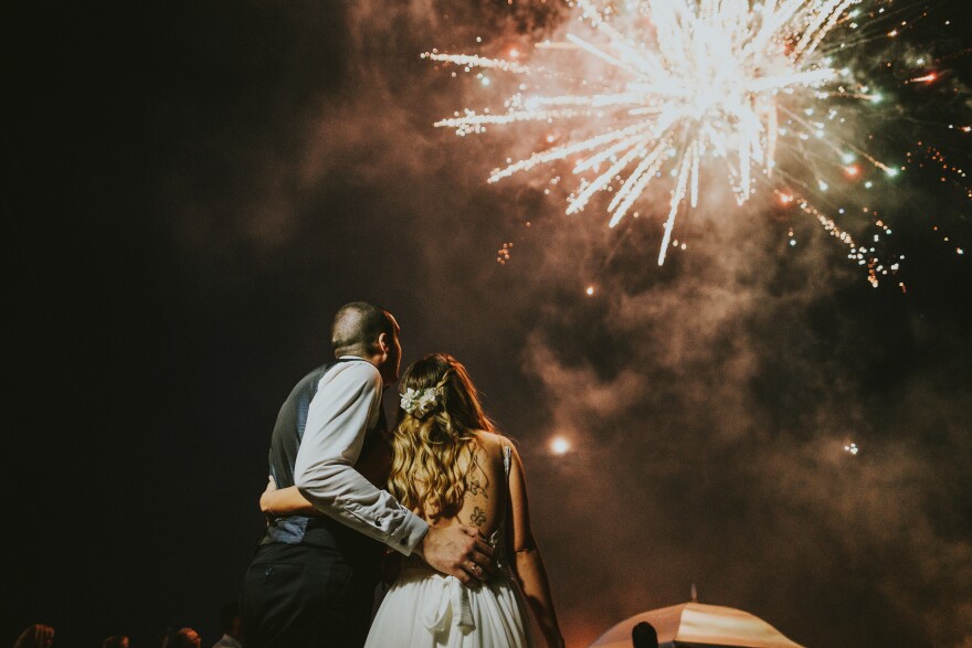 A man and a woman in wedding attire embrace one another. Above them, a firework explodes.