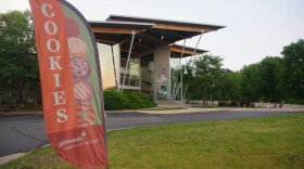 A photo of the Girl Scout Heart of Michigan, Kalamazoo Regional Center on June 17, 2024.  In the foreground is a flag style lawn banner that reads "cookies."  The word is spelled out in white letters on a light orange background with the letters stacked on top of each other vertically on the left side of the banner. On the right vertical side of the banner on a green background is a picture of five types of Girl Scout Cookies, stacked from the top to the bottom of the flag.  And at the bottom, in the lower quarter of the banner in white letters on a dark orange background it reads "Girl Scouts Heart of Michigan" in all lowercase letters.   Behind the flag is the driveway around the front of the concrete building with large panes of glass and a colorful mural on the concrete wall.  The building has  three distinct roof lines that are designed to loosely resemble a person with arms outstretched.  Bushes and trees are close to the building, on the building side of the drive.  A yellow-green lawn is in the foreground.