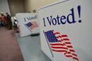 Cardboard voting dividers are shown sitting on tables with the words "I Voted!" printed in red, white and blue. In the background, some people can be seen standing.