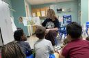 A woman is sits in a chair and reads a book to students in front of her. She props the book for them to see. The students are sitting with their backs facing the camera.