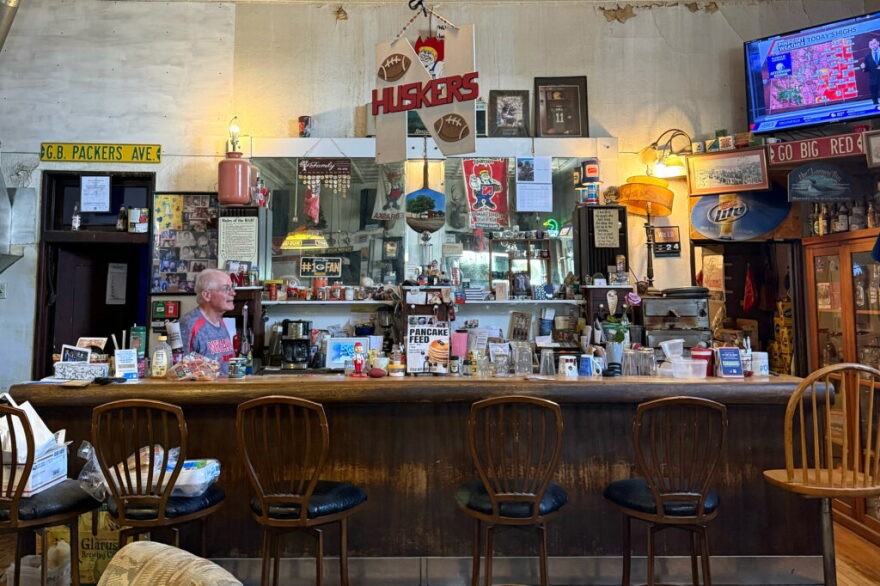 Behind a solid wood bar, photos, tchotchkes and glassware crowd shelves advertising a pancake feed and Nebraska Huskers. A television in the upper right corner shows the weather.