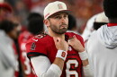 White football player with brown beard holds hands up to his chest wearing a red jersey with other football players blurred out in the photo behind them