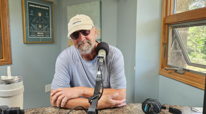 A bearded man in a blue shirt, sunglasses and baseball cap looks into the camera. He is sitting behind a microphone in a radio studio.