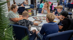 A group of five kids is sitting at a circular folding table with an adult eating chips and popcorn. They're playing the classic board game Clue.