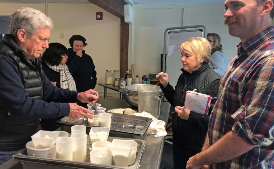 David Asher stands with students in his cheesemaking course. There is cheese on a table.
