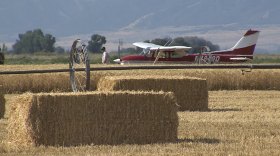 A small red private plane sits in a wheat field. A man stands in front of the plane and another man in a uniform stands farther away from the man closer to the plane.