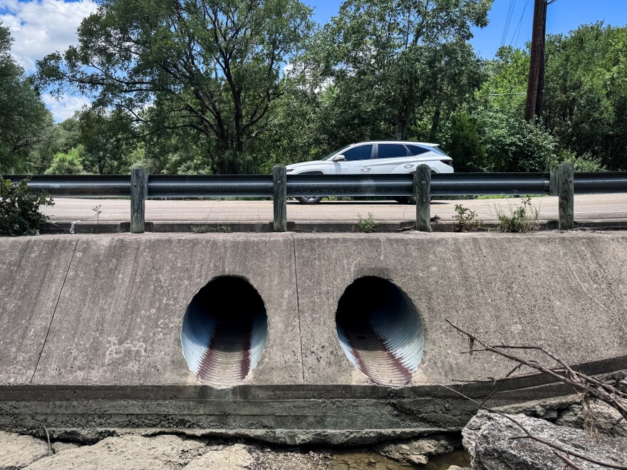 Two 36-inch culverts set in concrete run underneath a road. The creek bed is dry. A white crossover car is driving by, partly obscured by the guardrail.