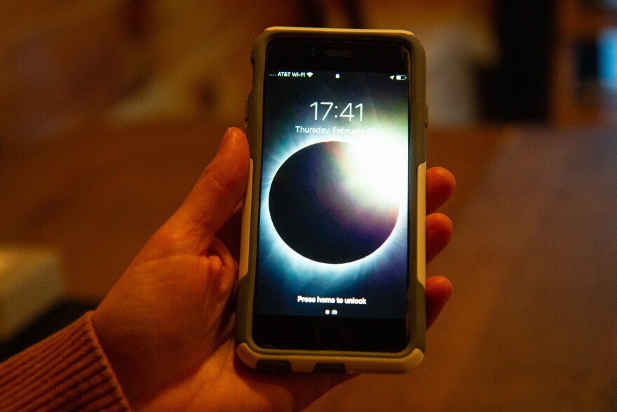 A photo of a person's hand holding up an iPhone with a background showing a total solar eclipse, the dark circle of the moon over the bright corona of the sun.