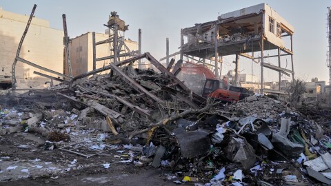 Rumble sits in a large piles with the frame of what's left of the facility seen behind it. A excavator sifts through the rubble of what used to be a building. 
