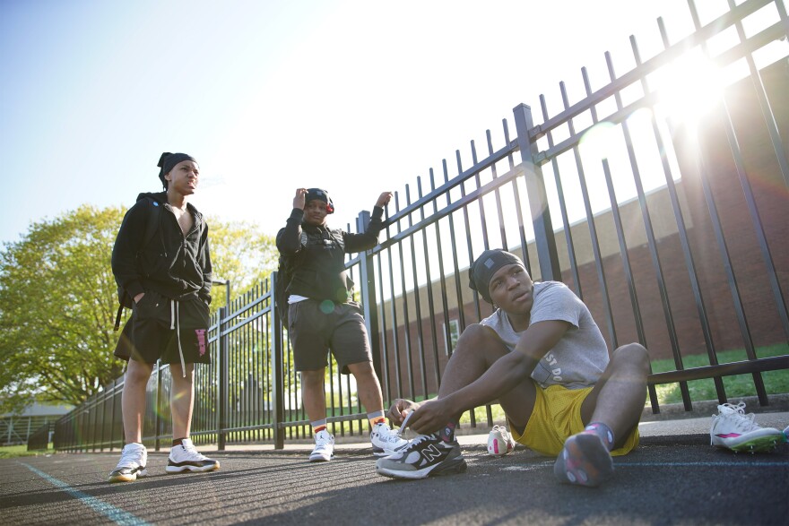 Two people in racing shoes stand by a fence as a person sitting on the ground ties his racing shoes.