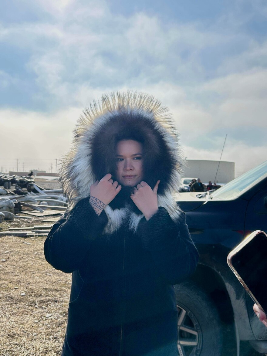A woman is dressed in a heavy parka with a fur-lined hood. She is posing for a portrait and holding the bottom of the hood with both hands. She is outdoors, a truck visible behind her and it is sunny with a cloudy sky.