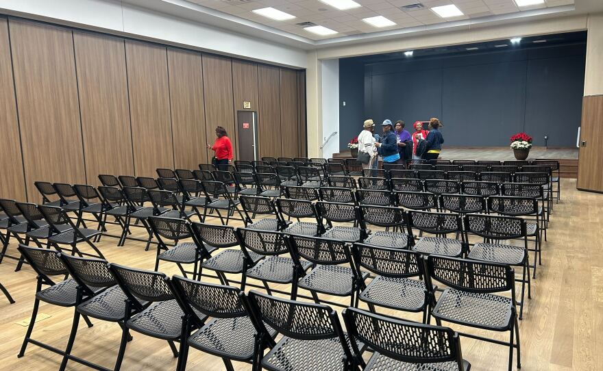 A room with wooden walls and floors, with rows of black chairs facing towards a stage, with a small group of people standing in front of it.