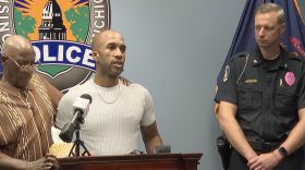 Two men stand at a podium and speak into a microphone next to a police officer