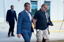 A man in a blue pin striped suit leads a man in handcuffs who is wearing a black t-shirt and cargo shorts off to the right. 