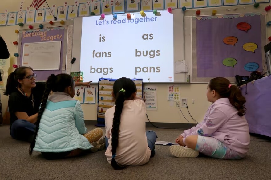 Three young students sit on the ground and look up toward a projected screen with their backs to the camera. Next to them, their teacher sits and points to the words on the board. It reads: "Let's read together: is, as, fans, bugs, bags, pans."