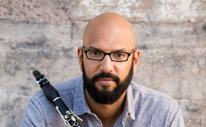 A bald man with a dark mustache and beard wearing glasses and blue button-down shirt. He's holding a clarinet.