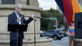 A man in a suit stands in front of a large concrete monument. He is speaking into a microphone and gesturing with his other hand. To his left an inflatable rainbow structure is visible, and a man in a rainbow shirt listens nearby.