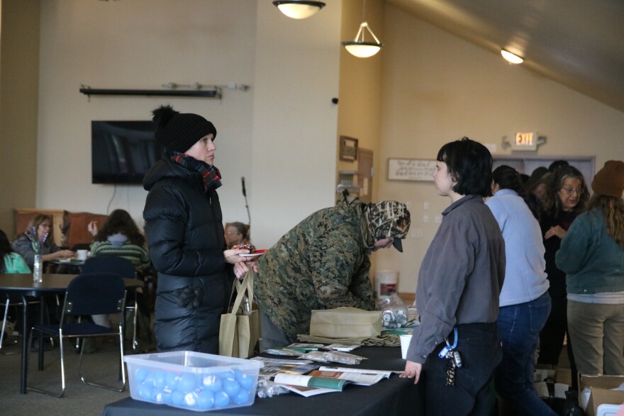 A woman in a gray long-sleeved shirt looks at a woman in a big black coat and black beanie and talks with her about county resources. There's stress balls and flyers on a table in front of them.