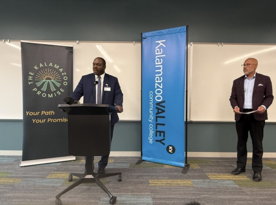 In a room with dark grey walls, a KPS Superintendent Darrin Slade stands at a black podium speaking in front of a large whiteboard on the wall. Behind him, two large standing banners read "The Kalamazoo Promise - Your path, your promise" and "Kalamazoo Valley community college". To the right of Slade, Kalamazoo Promise CEO Von Washington Jr. stands holding a sheet of paper. 